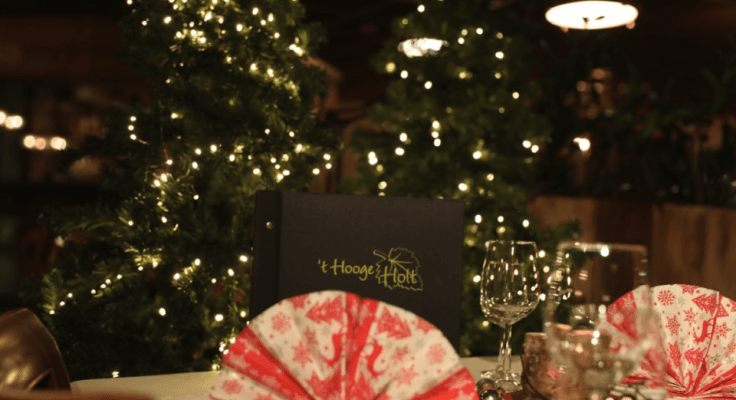 Een tafel met wijnglazen gevouwen servetten en een menukaart met 't Hooge Holt erop, met in de achtergrond twee kerstbomen met lichtjes